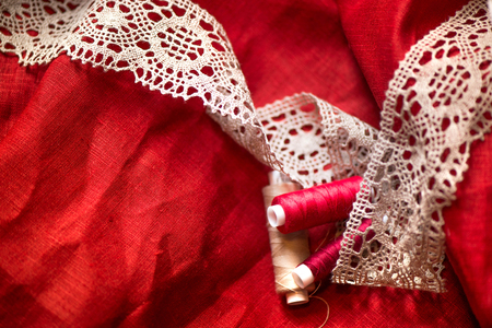 A macro shot of a red and white thread spools and a white, crochet natural linen lace trim on a dark red linen fabric, being used while sewing tablecloth by using a sewing machineの写真素材