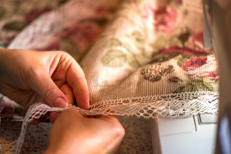 Making table decorations. A shot of woman sewing a natural beige linen tablecloth, towels and napkins with rose print and a crochet white linen lace trim, using a sewing machine and needlesの写真素材