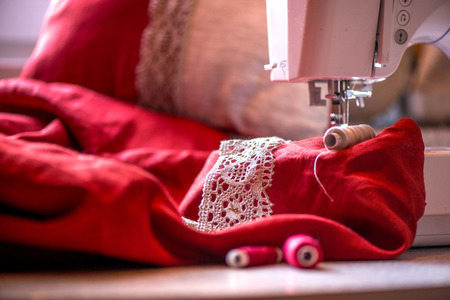 A macro shot of red and white thread spools and a white, crochet natural linen lace trim on a dark red linen fabric, being used while sewing tablecloth by using a sewing machineの写真素材