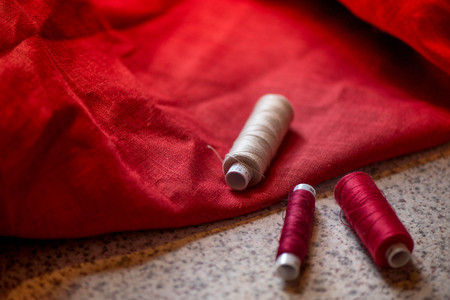 A macro shot of beige thread spool and a white, crochet natural linen lace trim on a dark red linen fabric, being used while sewing tablecloths, napkins and towels by using a sewing machine, needles.の写真素材