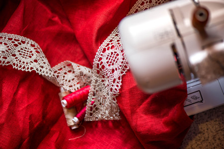 A macro shot of red and white thread spools and a white, crochet natural linen lace trim on a dark red linen fabric, being used while sewing tablecloth by using a sewing machineの写真素材
