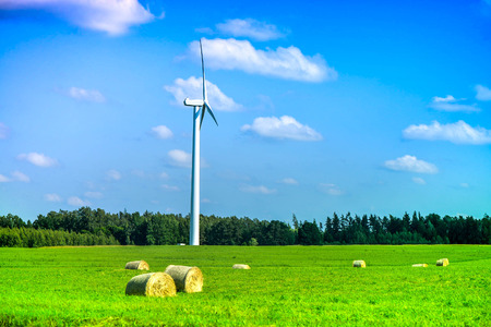 Summer scene of wind turbine in the middle of meadow full of straw bales, next to forest, during...