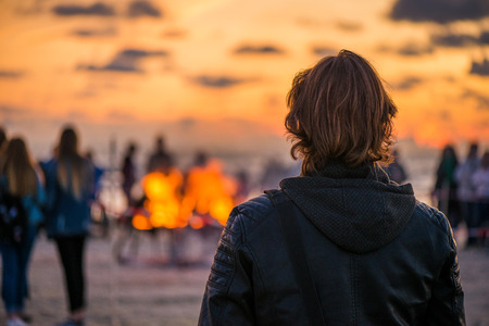Lonely young man looking at the sunset and waiting his love. Romantic bonfire night at seaside. People gathering together to celebrate Night of ancient lights. Large burning campfire with glowing flameの写真素材
