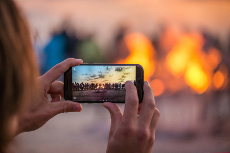 Woman is taking picture of sunset with smart phone. Romantic bonfire night at seaside. People gathering together to celebrate Night of ancient lights. Large burning campfire with soft glowing flame.の写真素材