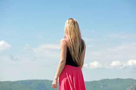Thoughtful young woman with long blonde hair and pink skirts standing at the edge of the mountain top, looking forward and dreaming of future.の写真素材