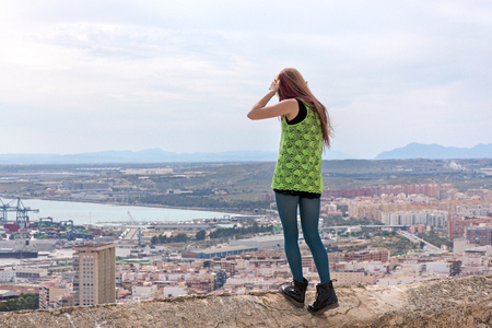 Young Caucasian girl standing on an edge of a Spanish conquistador fortress wall made of ancient stone and marble to enjoy the view over a modern metropolitan city. Girl ready to jump, suicide conceptの写真素材