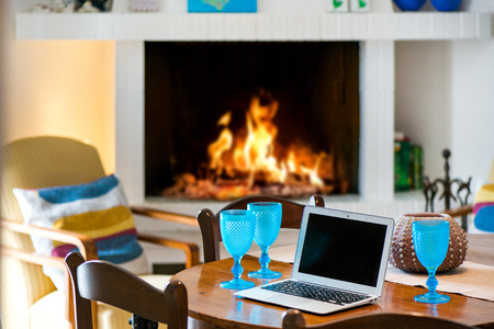 Laptop standing on ancient, vintage and old oak table in cozy living room next to fireplace with glowing, soft burning fire. Empty glasses of wine or beer. Waiting guests. Concept of working at homeの写真素材