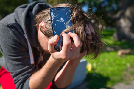 Broken and damaged smartphone with cracks on glass, screen. Young man is disappointed and sad, holding phone in hands. Concept of anger, rage and accident.の写真素材
