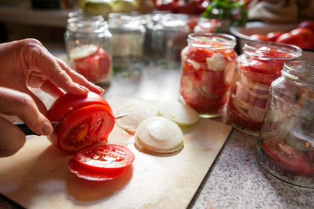 Canning fresh tomatoes with onions in jelly marinade. Woman hands putting red ripe tomato slices and onion rings in jars. Basil, parsley leaves on top of onions. Vegetable salads for winterの写真素材