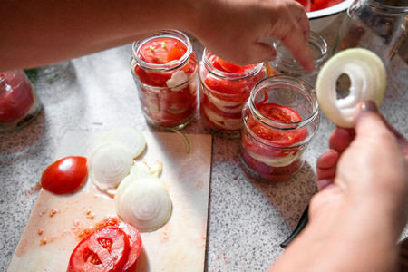 Canning fresh tomatoes with onions in jelly marinade. Woman hands putting red ripe tomato slices and onion rings in jars. Basil, parsley leaves on top of onions. Vegetable salads for winterの写真素材