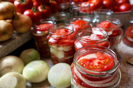 Canning fresh tomatoes with onions in jelly marinade. Woman hands putting red ripe tomato slices and onion rings in jars. Basil, parsley leaves on top of onions. Vegetable salads for winterの写真素材
