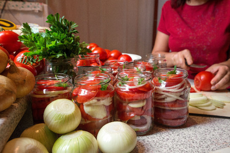 Canning fresh tomatoes with onions in jelly marinade. Woman hands putting red ripe tomato slices and onion rings in jars. Basil, parsley leaves on top of onions. Vegetable salads for winterの写真素材