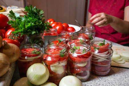 Canning fresh tomatoes with onions in jelly marinade. Woman hands putting red ripe tomato slices and onion rings in jars. Basil, parsley leaves on top of onions. Vegetable salads for winterの写真素材
