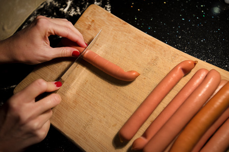 Preparing tanned wiener sausages in pizza dough. Cutting sausages on wooden board before baking. Dark background in kitchen. Making snacks for family and friendsの写真素材