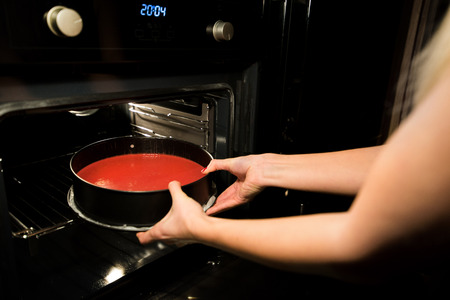 Woman is cooking in kitchen, baking a red velvet cake. Taking out or putting a biscuit tray filled with dough in oven. Delicious, homemade dessertの写真素材