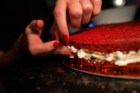 Woman spreading whipped cream with fingers inside freshly made cake. Beautiful home made red velvet cake decorated with whipped cream and raspberry crumbs. Concept of anti sanitary cookingの写真素材