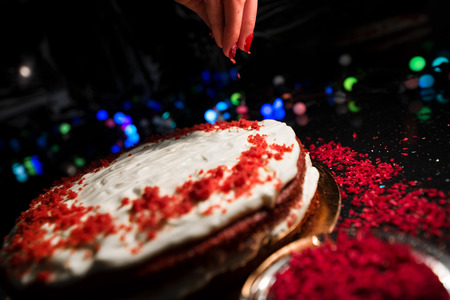 Woman sprinkling raspberry crumbs, icing sugar over freshly made cake, creating ornaments and decorating dessert. Beautiful home made red velvet cake decorated with whipped cream and raspberry crumbsの写真素材