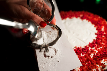 Woman hands sprinkling icing sugar over freshly made cake, creating ornaments and decorating dessert. Beautiful home made red velvet cake decorated with whipped cream and raspberry crumbs.の写真素材