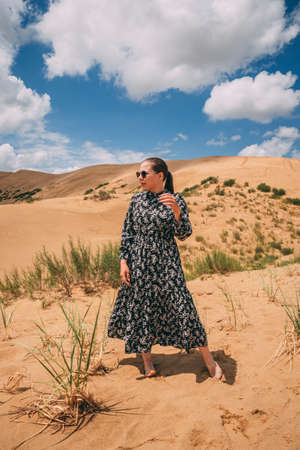 Beautiful landscape in sand dunes. A girl in a pink light dress walks through the desert against the blue sky. Safari in Kazakhstan. Golden Sands of Mangystau at sunsetの写真素材