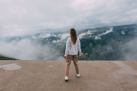 A young traveling woman stands on the edge of a mountain cliff and watching a beautiful view of the forest, mountains and clouds on vacation. The woman travels alone. Dagestan mountainsの写真素材