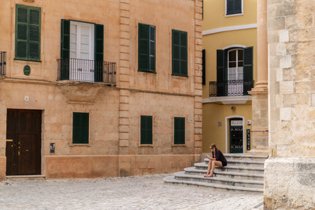 Ciutadella, Menorca / Spain; 10 05 2015: Beautiful square with large stone facades and dark green wooden shutters.In the right corner the stairs of the entrance to a church, with a woman sitting onのeditorial素材