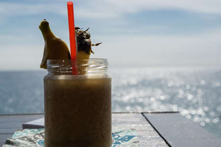 Natural milkshake with pineapple and banana in a glass jar and red straw, on a wooden table facing...
