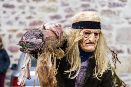 Ituren, Navarre, Spain; 01 27 2020: Person in old clothes, long blond wig fastened with ribbon and mask of a sad old man with a cigarette in his nose carrying a log on his shoulder during the carnivalのeditorial素材