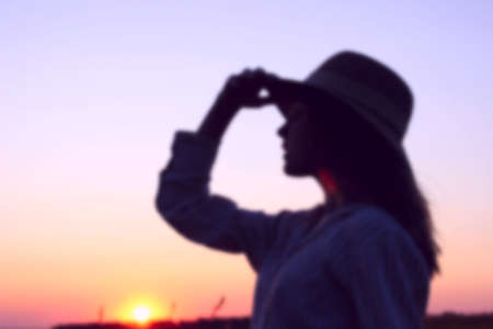 Blurry silhouette of teenage girl wearing a sun hat and looking to the left, close up portait, space for text. Female portrait, sunset background, cropped shot. People, travel concept.の写真素材