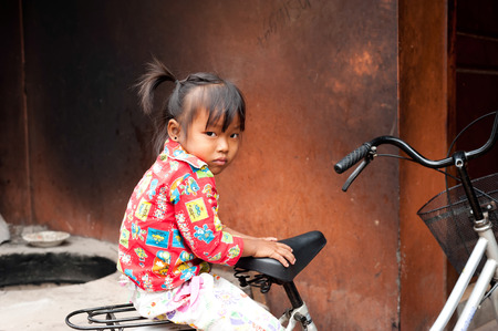 SIEM REAP, CAMBODIA - DEC 22, 2013: Unidentified khmer cute little girl in colorful clothes sitting on  bicycle at street over grunge wall background on Dec 22, 2013 in Siem Reap, Cambodiaのeditorial素材