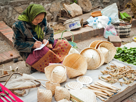 LUANG PRABANG, LAOS - DEC 8: Unidentified woman selling traditional asian conical hats, wooden kitchen utensils and other bamboo handcrafts at marketplace on Dec 8, 2013 in Luang Prabang, Laosのeditorial素材