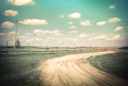 Sunny day in countryside. Summer landscape with rural road, wooden fence and electric poles under blue cloudy sky. Nature background in vintage styleの写真素材