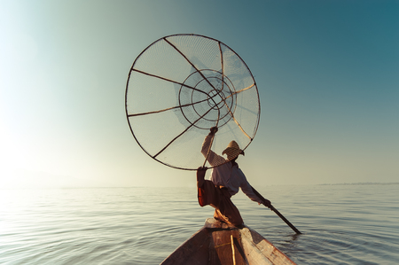 Burmese fisherman on bamboo boat catching fish in traditional way with handmade net. Inle lake, Myanmar (Burma) travel destinationの写真素材