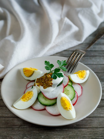 Plate with healthy spring vegetarian salad with radish, cucumber, egg and parsley with sour cream. and mustard, top view on wooden background with fork and napkin, russian summer food. Verticalの写真素材