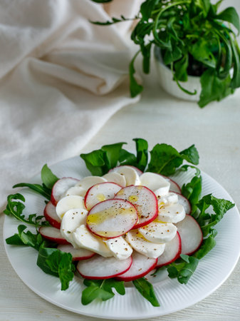Plate with healthy spring vegetarian salad with radish, mozzarella cheese and rucola with olive oil. top view on white wooden background with napkin, summer food. Verticalの写真素材