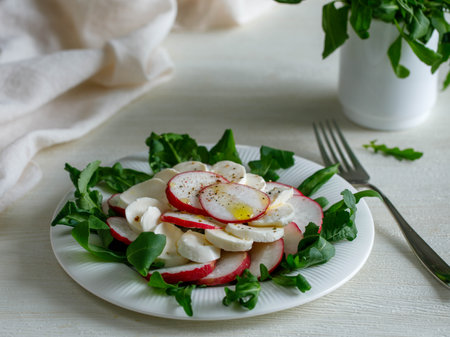 Plate with healthy spring vegetarian salad with radish, mozzarella cheese and rucola with olive oil. top view on white wooden background with napkin, summer foodの写真素材