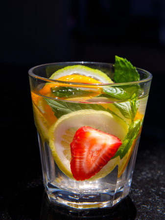 Citrus and mint leaves infused water, cocktail, lemonade in glass. Summer iced cold drink with lemon, lime, orange, strawberry, mint on black marble background, close uo, side view, verticalの写真素材