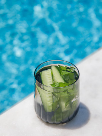 Cucumber and mint leaves infused water, cocktail, lemonade in glass near pool. Summer iced cold drink with cucumber, ice and mint on blue water white marble background, copy space, top view, verticalの写真素材