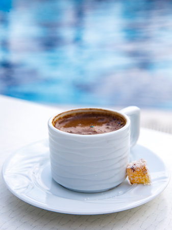 Delicious traditional Turkish Coffee served with Turkish Delight lokum in traditional white ceramic cup on white background in sunny day at swimming pool, close up verticalの写真素材