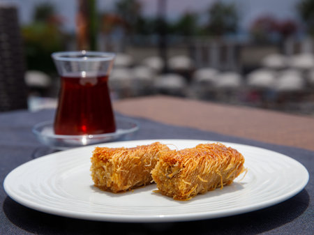 Pistachio kadayif on white plate with turkish tea on table in outdoor cafe. Traditional Middle Eastern Flavors. The local name of kadayif Fistikli kadayif. Side view, close up, hard shadowsの写真素材