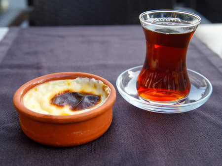 Baked rice pudding turkish milky dessert sutlac in earthenware casserole with turkish tea on black cloth on table, close upの写真素材