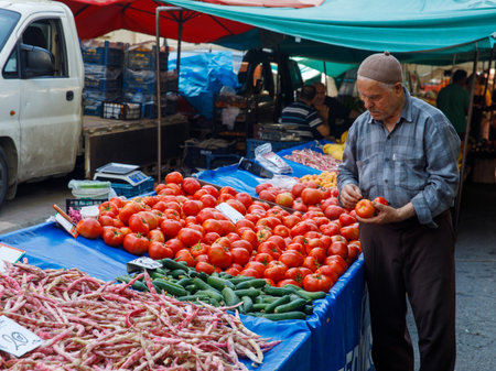 Elderly buyer with gray hair and traditional hat buying tomatoes at weekly local farmer market. He carefully choosing vegetables. There are lots of different ripe vegetables. Alanya, Turkey 06june2022のeditorial素材