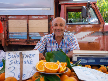 Elderly seller with gray hair sells orange fruit at weekly local farmer market. He smiles. There are many whole and sliced oranges in foreground and old truck in background. Alanya, Turkey 06june2022のeditorial素材