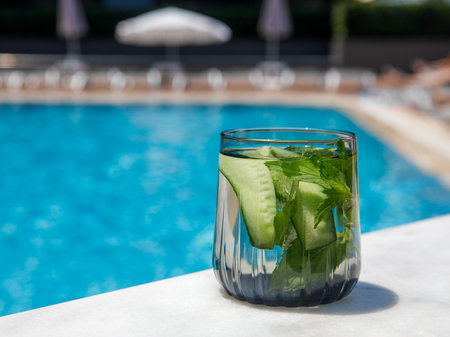 Cucumber and mint leaves infused water, cocktail, lemonade in glass near pool. Summer iced cold drink with cucumber, ice and mint on blue water and white marble background, copy space, side viewの写真素材