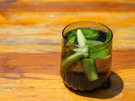Cucumber and mint leaves infused water, cocktail, lemonade in glass in cafe. Summer iced cold drink with cucumber, ice and mint on orange wooden table outside background, copy space, side view, sunnyの写真素材