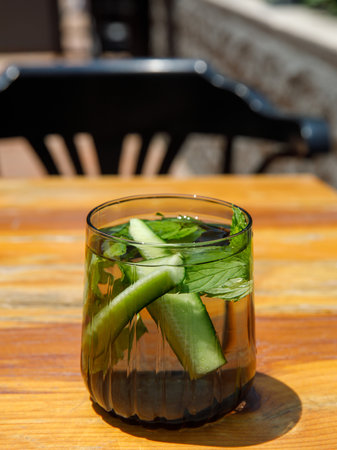 Cucumber and mint leaves infused water, cocktail, lemonade in glass in cafe. Summer iced cold drink with cucumber, ice and mint on orange wooden table outside background, copy space, vertical, sunnyの写真素材