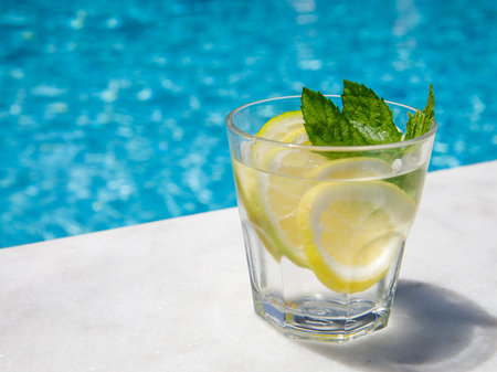 Citrus and mint leaves infused water, cocktail, lemonade in glass near pool. Summer iced cold drink with lemon, lime, ice and mint on blue water and white marble background, copy space, side viewの写真素材