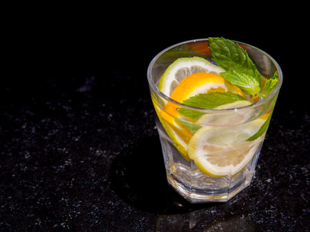 Citrus and mint leaves infused water, cocktail, lemonade in glass. Summer iced cold drink with lemon, lime, orange, mint on black marble background, copy space, side viewの写真素材