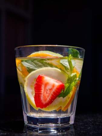 Citrus and mint leaves infused water, cocktail, lemonade in glass. Summer iced cold drink with lemon, lime, orange, strawberry, mint on black marble background, copy space, side view, verticalの写真素材