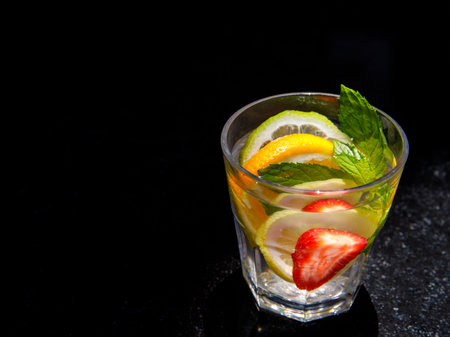Citrus and mint leaves infused water, cocktail, lemonade in glass. Summer iced cold drink with lemon, lime, orange, strawberry, mint on black marble background, copy space, side viewの写真素材