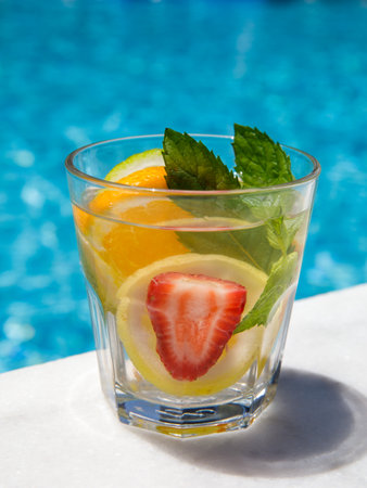 Citrus mint leaves infused water, cocktail, lemonade in glass near pool. Summer iced cold drink with lemon, lime, orange, strawberry, mint on blue water white marble background, vertical, side viewの写真素材